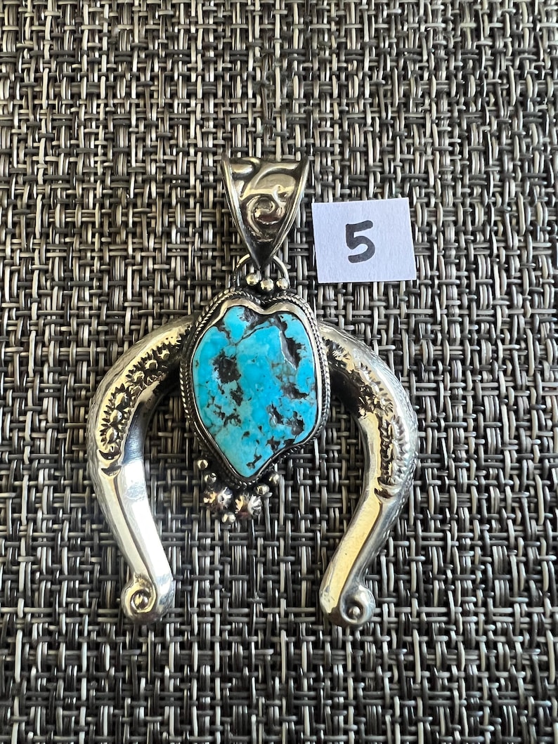 May include: A silver pendant featuring a central irregular turquoise stone with black matrix, set within a horseshoe-shaped frame with engraved floral patterns and small silver beads. The bail has an etched design and a white tag with the number 5.