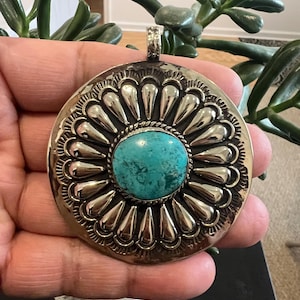 May include: A circular silver pendant featuring a central round turquoise stone surrounded by a twisted wire border and a radiating pattern of smooth, teardrop-shaped silver petals. The pendant includes a small silver bail at the top.