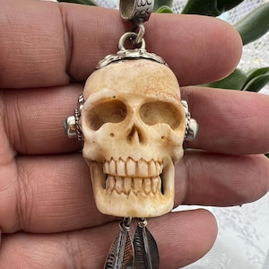 May include: A cream-coloured skull pendant with detailed carving, silver accents, and small turquoise stones. Two silver feather charms hang below the skull. The pendant has a decorative bail for attaching to a chain.