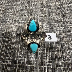 May include: A silver ring featuring two teardrop-shaped turquoise stones, one at the top and one at the bottom, accented by three metal flowers in the center. The silver setting includes leaf-like engravings and twisted wire details. A small white tag with the number 3 is visible.