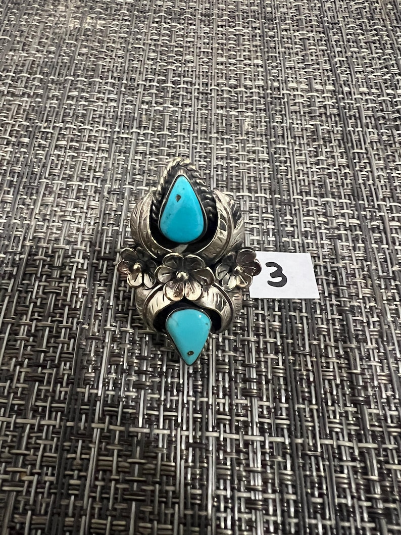 May include: A silver ring featuring two teardrop-shaped turquoise stones, one at the top and one at the bottom, accented by three metal flowers in the center. The silver setting includes leaf-like engravings and twisted wire details. A small white tag with the number 3 is visible.