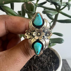 May include: A silver ring featuring two teardrop-shaped turquoise stones set vertically. The center of the ring is decorated with three small silver flowers surrounded by engraved leaf patterns. The metal has a polished finish with dark oxidation.