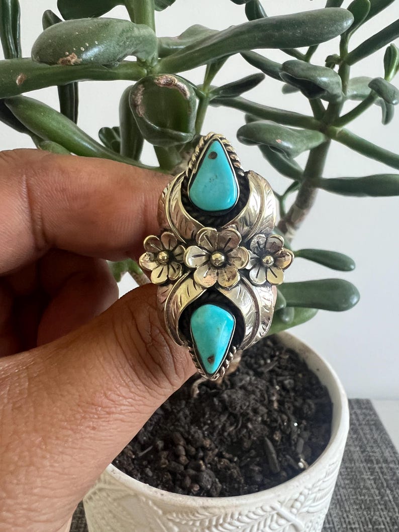May include: A silver ring featuring two teardrop-shaped turquoise stones set vertically. The center of the ring is decorated with three small silver flowers surrounded by engraved leaf patterns. The metal has a polished finish with dark oxidation.