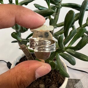 May include: A silver-toned metal ring featuring an open, adjustable band design with intricate, embossed scrollwork patterns along the outer surface. The ring has a flat, rounded top section and a polished finish, showcasing detailed engravings.