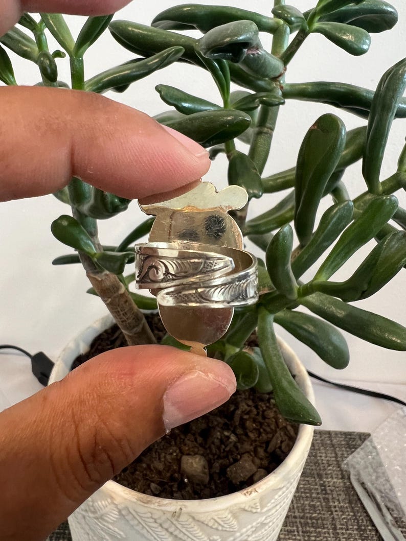 May include: A silver-toned metal ring featuring an open, adjustable band design with intricate, embossed scrollwork patterns along the outer surface. The ring has a flat, rounded top section and a polished finish, showcasing detailed engravings.