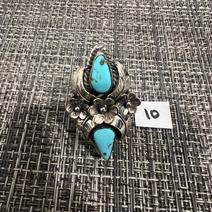 May include: A silver ring featuring two teardrop-shaped turquoise stones, decorative metal leaves, and three small metal flowers. The ring is positioned next to a small white paper tag with the handwritten number 10 in black ink.