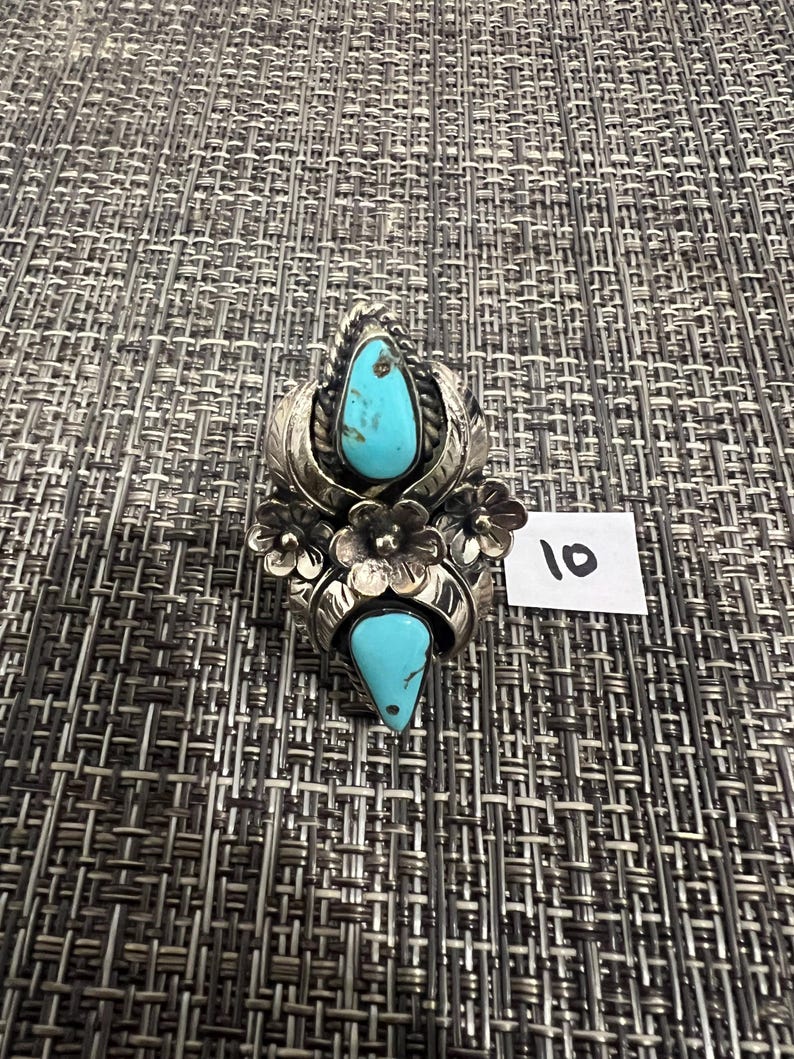 May include: A silver ring featuring two teardrop-shaped turquoise stones, decorative metal leaves, and three small metal flowers. The ring is positioned next to a small white paper tag with the handwritten number 10 in black ink.