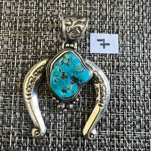 May include: A silver pendant featuring a central, irregular turquoise stone with dark brown matrix veins. The pendant has a horseshoe-shaped frame with engraved patterns and a decorative bail at the top. A small white paper tag with the number 7 is visible.