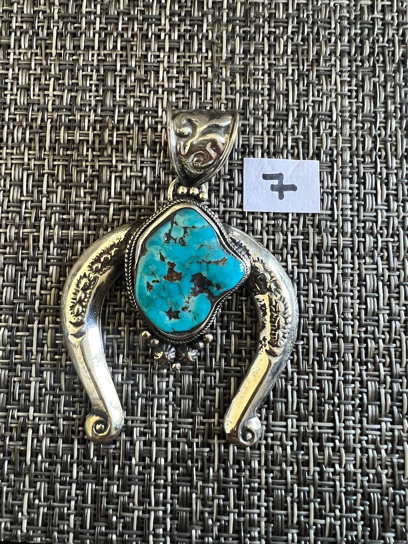 May include: A silver pendant featuring a central, irregular turquoise stone with dark brown matrix veins. The pendant has a horseshoe-shaped frame with engraved patterns and a decorative bail at the top. A small white paper tag with the number 7 is visible.