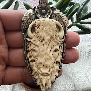 May include: A detailed pendant featuring a carved bison head in cream and brown tones, set within a silver-coloured frame. The pendant has a floral accent at the top and is designed with intricate details, showcasing a rustic, handcrafted aesthetic.