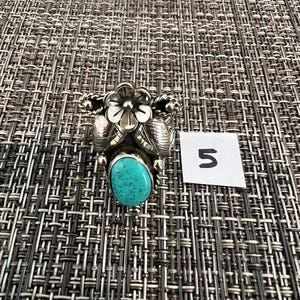 May include: A silver ring featuring a central oval turquoise stone, accented by a metal flower and leaf design. The ring is positioned next to a small white square paper with the handwritten number 5 in black ink, showcasing detailed metalwork.