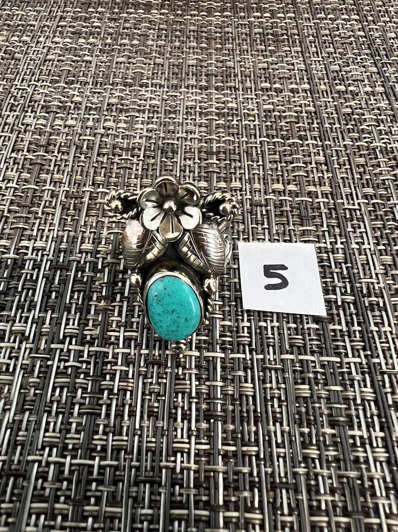 May include: A silver ring featuring a central oval turquoise stone, accented by a metal flower and leaf design. The ring is positioned next to a small white square paper with the handwritten number 5 in black ink, showcasing detailed metalwork.