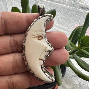 May include: Crescent moon pendant with a carved face, set in a silver-toned metal frame. The moon has a cream-colored surface with detailed facial features. The pendant is suspended from a small loop at the top.