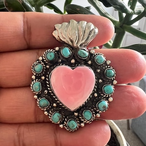 May include: A heart-shaped pendant featuring a smooth pink center stone surrounded by a border of small, round turquoise stones set in oxidized silver. The top of the pendant is adorned with a silver flame-like design with textured detailing.
