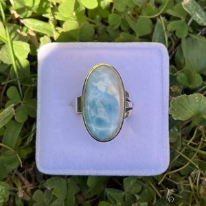 Op de afbeelding: Een zilveren ring met een grote ovale blauwe en witte edelsteen. De ring ligt in een wit fluwelen doosje.