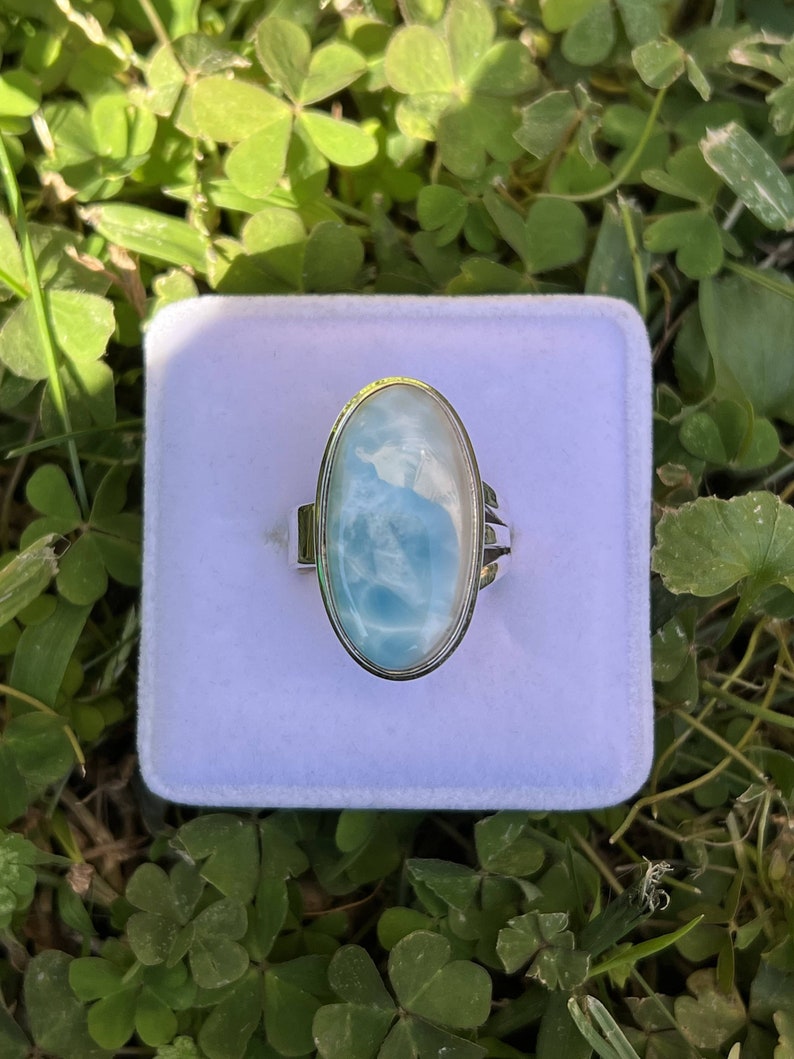 Op de afbeelding: Een zilveren ring met een grote ovale blauwe en witte edelsteen. De ring ligt in een wit fluwelen doosje.