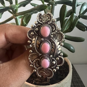 Op de afbeelding: Een grote zilveren ring met drie verticaal geplaatste, ovale roze cabochonstenen. De zilveren zetting is rijk versierd met gestempelde bladmotieven, gedraaide draadranden en kleine zilveren bolletjes rondom de stenen.