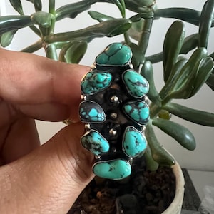 May include: A statement ring featuring seven irregular, polished turquoise stones with dark matrix veining, set in a dark oxidized metal base. The stones are arranged in a vertical cluster with small silver bead accents between each turquoise piece.