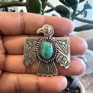 Op de afbeelding: Een zilverkleurige broche in de vorm van een gestileerde vogel met gespreide vleugels. Het midden bevat een ovale turkoois steen omringd door een kralenrand. De vleugels en staart zijn versierd met gegraveerde geometrische patronen.