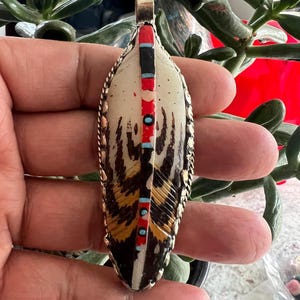 May include: A feather-shaped pendant with a silver-colored metal frame. The pendant features a cream-colored background with brown and black feather details. It has red, black, and turquoise accents. The pendant is held in a hand.