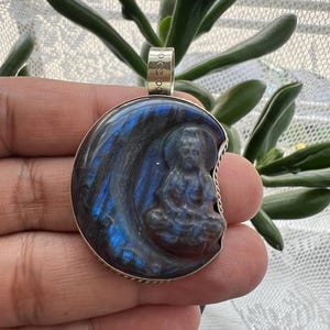 May include: A silver pendant with a carved image of a seated figure within a crescent moon shape. The pendant features blue and black iridescent stone, and a silver bail for a chain. The pendant is held in a hand.