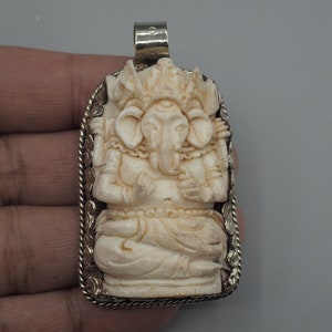 Puede incluir: Un colgante de marfil blanco tallado de la deidad hindú Ganesha, con un marco de plata y un bucle para colgar.
