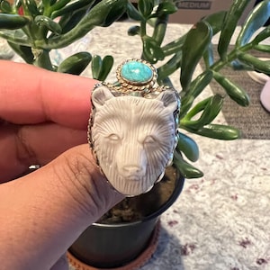 Op de afbeelding: Zilveren ring met een gebeeldhouwde witte berenkop. De ring heeft een goudkleurige zetting en een turkooizen steen. De ring wordt voor een groene plant gehouden.