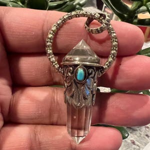 May include: A clear quartz crystal pendant with a silver-coloured metal setting. The pendant features a turquoise-coloured stone and decorative feather-like details. The crystal is approximately 5 cm long and is suspended from a circular bail.