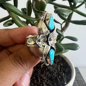 May include: A silver ring featuring two teardrop-shaped turquoise stones, two embossed metal flowers, and engraved leaf patterns. The band is wide and decorated with intricate scrollwork, showcasing a polished finish with oxidized detailing.