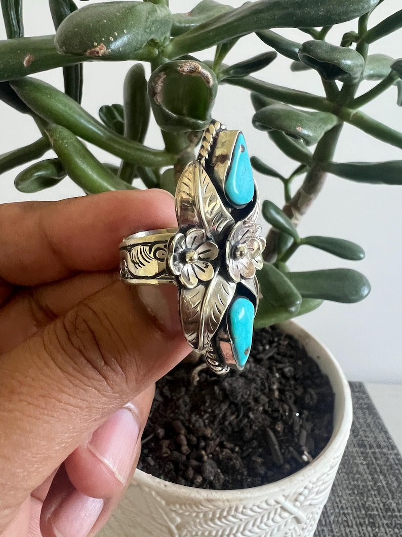 May include: A silver ring featuring two teardrop-shaped turquoise stones, two embossed metal flowers, and engraved leaf patterns. The band is wide and decorated with intricate scrollwork, showcasing a polished finish with oxidized detailing.