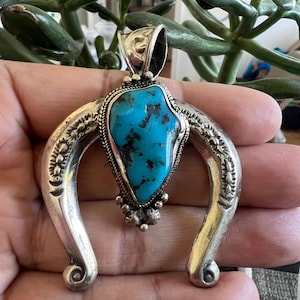 Puede incluir: Un colgante de plata en forma de herradura con una piedra turquesa central de color azul brillante irregular con inclusiones de matriz oscura. El marco de plata está decorado con patrones estampados, un borde de alambre trenzado y pequeñas esferas de plata.