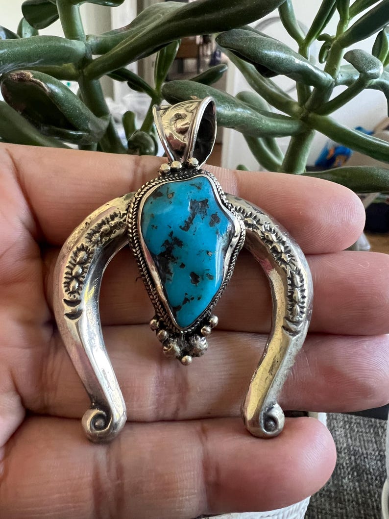 May include: A horseshoe-shaped silver pendant featuring a central, irregular, bright blue turquoise stone with dark matrix inclusions. The silver frame is decorated with stamped patterns, a twisted wire border, and small silver spheres at the base.