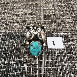 May include: A silver ring featuring a central oval turquoise stone with dark matrix patterns, topped with a detailed five-petal flower and two embossed leaves. The metal has a polished finish with decorative twisted wire accents on the sides.
