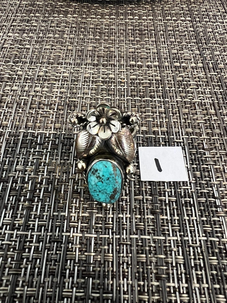May include: A silver ring featuring a central oval turquoise stone with dark matrix patterns, topped with a detailed five-petal flower and two embossed leaves. The metal has a polished finish with decorative twisted wire accents on the sides.