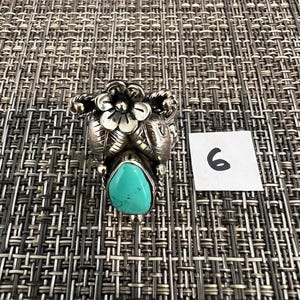 May include: A silver ring featuring a teardrop-shaped turquoise stone at the base, topped with a detailed silver flower and leaf design. The metal has a polished finish with darkened recessed areas. A small white square with the number 6 is visible.