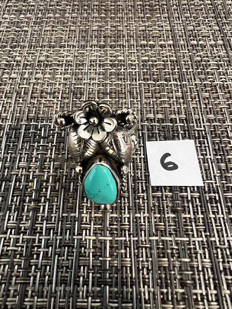 May include: A silver ring featuring a teardrop-shaped turquoise stone at the base, topped with a detailed silver flower and leaf design. The metal has a polished finish with darkened recessed areas. A small white square with the number 6 is visible.