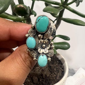May include: A silver ring featuring three oval turquoise stones, a central metal flower, and decorative scrollwork. The design includes a polished silver leaf element and small silver spheres arranged around the stones and floral centerpiece.