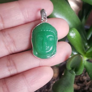 May include: A green stone pendant carved into the shape of a Buddha head, set within a simple silver-toned metal frame. The pendant features a small silver-toned metal bail at the top for attachment to a chain or cord, showcasing smooth surfaces.
