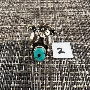 May include: A silver ring featuring a central oval turquoise stone with a black inclusion, topped with a detailed silver flower and leaf design. A small white square tag with the handwritten number 2 is positioned next to the ring.