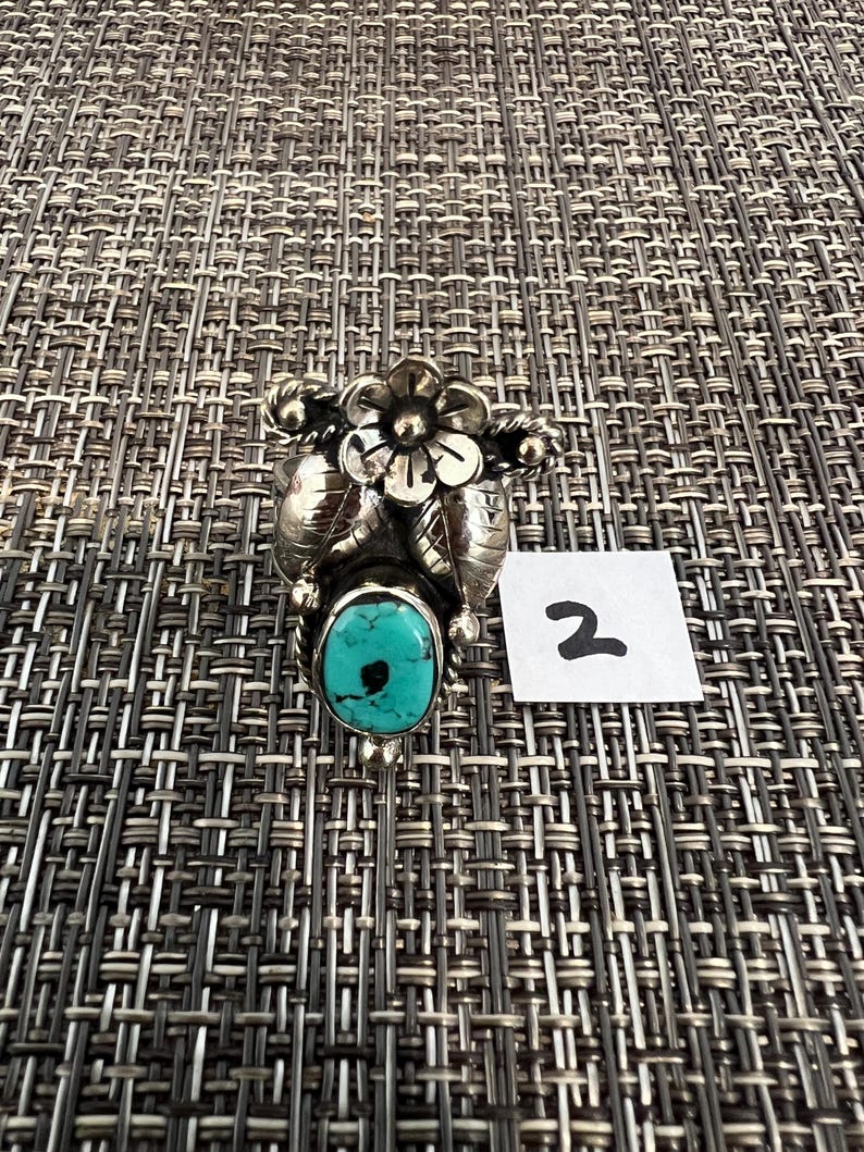 May include: A silver ring featuring a central oval turquoise stone with a black inclusion, topped with a detailed silver flower and leaf design. A small white square tag with the handwritten number 2 is positioned next to the ring.
