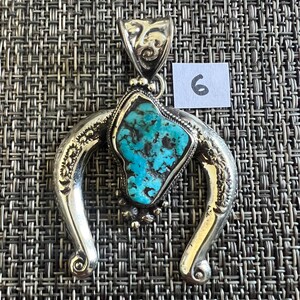 May include: A silver pendant featuring a central irregular turquoise stone with dark matrix veins, set within a polished horseshoe-shaped frame with engraved details and a decorative bail. A small white square tag with the number 6 is visible.