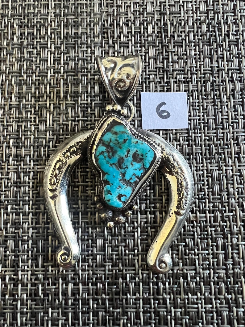 May include: A silver pendant featuring a central irregular turquoise stone with dark matrix veins, set within a polished horseshoe-shaped frame with engraved details and a decorative bail. A small white square tag with the number 6 is visible.
