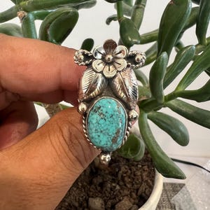 May include: A silver ring featuring a central oval turquoise stone with dark matrix patterns. The setting includes a polished silver flower at the top, two etched silver leaves below the flower, and decorative twisted wire and silver bead accents.