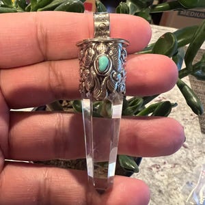 Może przedstawiać: Przezroczysty wisiorek z kryształu kwarcu ze srebrną metalową nasadką i krawatem. Nasadka ma ozdobne detale i mały, okrągły turkusowy kamień. Kryształ jest fasetowany i zwęża się do punktu.