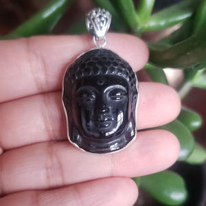 Puede incluir: Un colgante de Buda de obsidiana negra con una argolla de plata adornada. El colgante está tallado con detalles intrincados y tiene un acabado pulido.