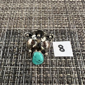 May include: A silver ring featuring a central teardrop-shaped turquoise stone with black matrix, accented by a sculpted metal flower and leaf design. A small white square tag with the handwritten number 8 is positioned next to the ring.