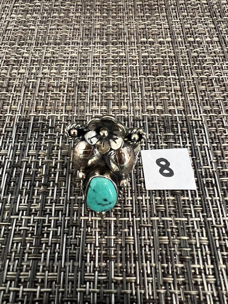 May include: A silver ring featuring a central teardrop-shaped turquoise stone with black matrix, accented by a sculpted metal flower and leaf design. A small white square tag with the handwritten number 8 is positioned next to the ring.