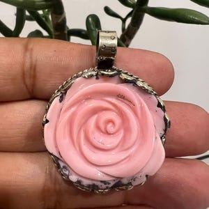 May include: A round pendant featuring a carved light pink rose set within a dark, textured metal frame. The pendant includes a silver-toned metal bail at the top for attachment, showcasing intricate detailing on the rose petals and the metal border.