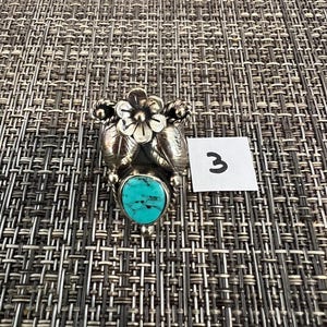 May include: A silver ring featuring a central oval turquoise stone with black matrix veining, topped with a detailed silver flower and two silver leaves. A small white square tag with the handwritten number 3 is placed next to the ring.