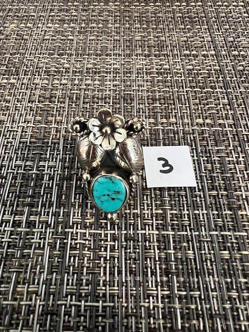 May include: A silver ring featuring a central oval turquoise stone with black matrix veining, topped with a detailed silver flower and two silver leaves. A small white square tag with the handwritten number 3 is placed next to the ring.
